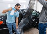 Male worker with spray wetting car tinting, tuning service. Mechanic applying vinyl tint on vehicle window in garage, tinted automobile glass