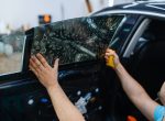 Male worker installs wetted car tinting, tuning service. Mechanic applying vinyl tint on vehicle window in garage, tinted automobile glass